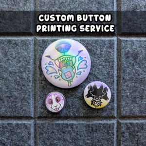 May include: Three custom-printed buttons with colorful designs. The largest button features a cartoon monster with a heart and a patch. The other two buttons have a bunny and a three-headed dog design. The text "CUSTOM BUTTON PRINTING SERVICE" is at the top.