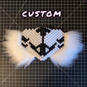 May include: A black and white beaded heart-shaped mask with white faux fur trim. The mask is on a black grid background. The word "CUSTOM" is written in pink above the mask.