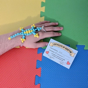 May include: A certificate of adoption for a pet named "Minasaurus" with a blue and yellow beaded lizard on a person's wrist. The certificate states that the person adopted the pet on April 12, 2024 and promises to take the pet on 10,000 fun adventures.