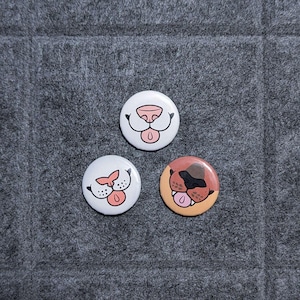 May include: Three round buttons with dog face designs. One button has a white background with a pink nose and tongue. Another has a white background with a pink nose and tongue. The third button has a brown and tan background with a pink tongue.