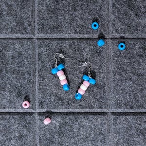 May include: A pair of handmade earrings featuring a transgender pride flag design. The earrings are made with blue, white, and pink beads. Several loose beads in matching colors are scattered around the earrings on a gray surface.