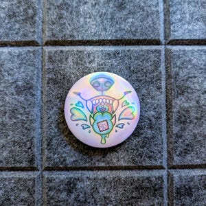 May include: A circular, iridescent badge featuring a cartoon dog-like face. The design includes a dog's nose, teeth, and a heart-shaped tongue, with blue, green, and pink accents. The badge is set against a grey, textured background.