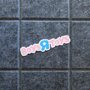 May include: A sticker with the text "Boys Я Toys" in pink and blue, with a star in the letter "Я". The sticker is on a gray, textured surface.