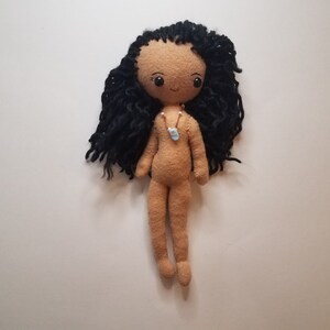 May include: A handmade doll with long black yarn hair, brown eyes, and a light brown felt body. The doll is wearing a simple necklace with a blue pendant.