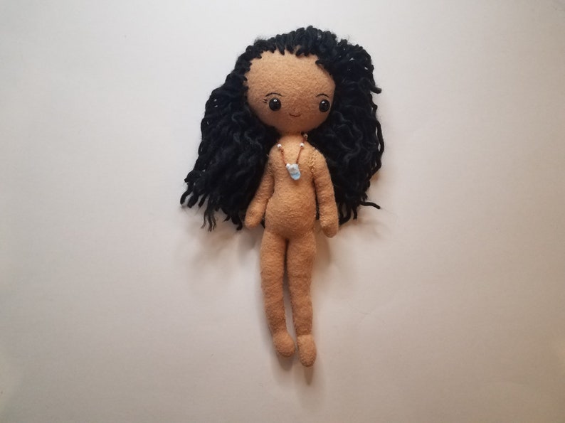 May include: A handmade doll with long black yarn hair, brown eyes, and a light brown felt body. The doll is wearing a simple necklace with a blue pendant.