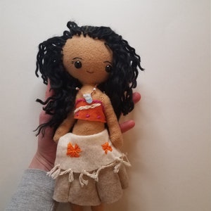 May include: Handmade felt doll with long black curly hair, brown eyes, and a brown face. She is wearing a white and orange top, a white skirt with orange flowers, and a white necklace with a blue pendant.