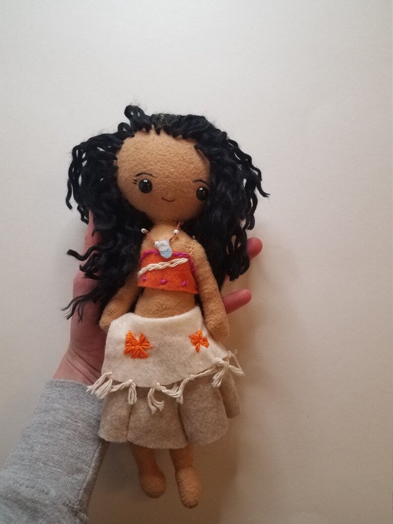 May include: Handmade felt doll with long black curly hair, brown eyes, and a brown face. She is wearing a white and orange top, a white skirt with orange flowers, and a white necklace with a blue pendant.