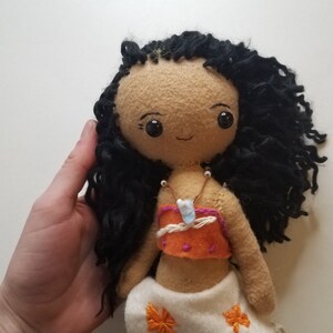 May include: Handmade doll with brown skin, black curly hair, and a brown and orange top. The doll is wearing a white skirt with floral embroidery and a necklace with a blue stone pendant.