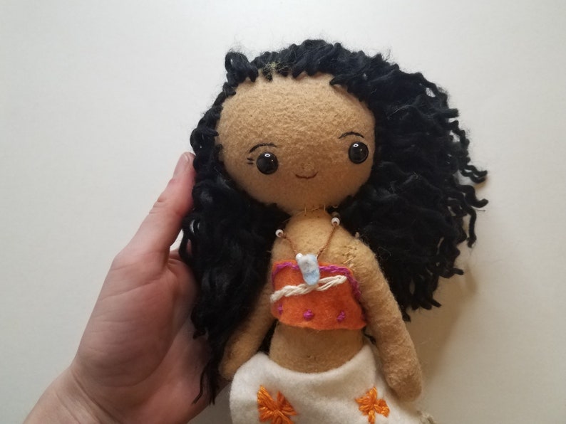May include: Handmade doll with brown skin, black curly hair, and a brown and orange top. The doll is wearing a white skirt with floral embroidery and a necklace with a blue stone pendant.