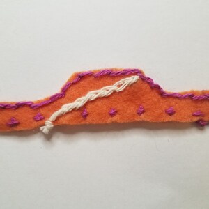May include: Orange felt applique with pink stitching and a white embroidered design. The applique is shaped like a car and has a simple design.