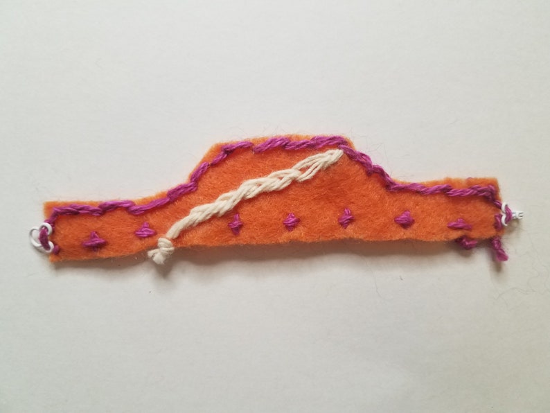 May include: Orange felt applique with pink stitching and a white embroidered design. The applique is shaped like a car and has a simple design.