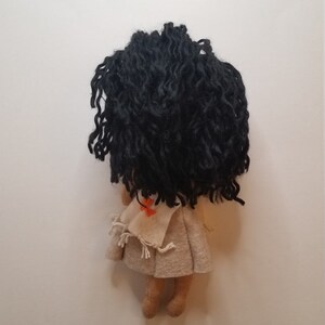 May include: A felt doll with long black curly hair, wearing a beige dress with a red and orange embroidered design. The doll is standing on a white background.