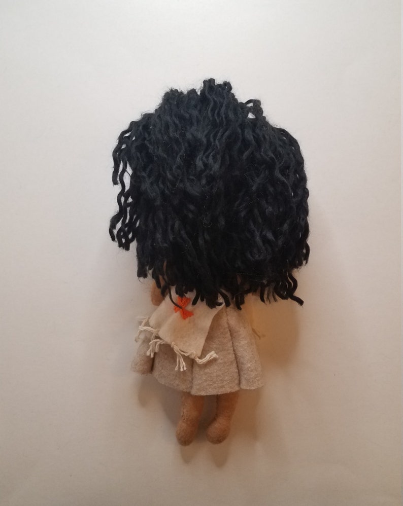 May include: A felt doll with long black curly hair, wearing a beige dress with a red and orange embroidered design. The doll is standing on a white background.