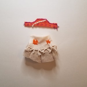 May include: A handmade doll outfit with a tan skirt with orange embroidery and a pink and orange headband.