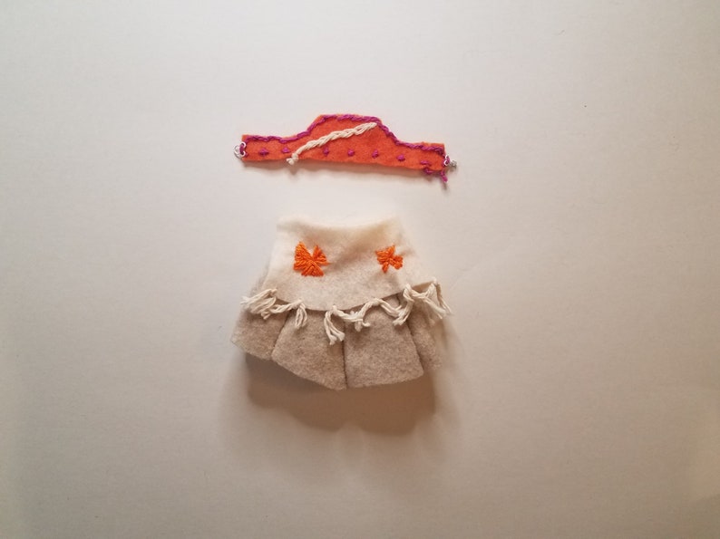 May include: A handmade doll outfit with a tan skirt with orange embroidery and a pink and orange headband.