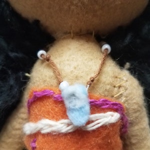 May include: A brown felt doll with black hair and a purple and orange felt outfit. The doll has white beads strung on a brown cord around its neck.