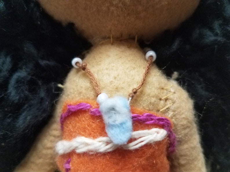 May include: A brown felt doll with black hair and a purple and orange felt outfit. The doll has white beads strung on a brown cord around its neck.
