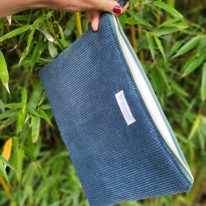 Peut inclure: Une pochette zippée en velours côtelé bleu sarcelle foncé avec une surface texturée à côtes et une petite étiquette en tissu blanc rectangulaire avec du texte noir cousue sur le devant. La pochette possède une fermeture éclair dorée.