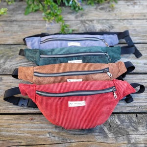 Puede incluir: Cinco riñoneras de pana en diferentes colores: rojo, marrón, verde, gris y azul. Cada riñonera tiene una cremallera y una pequeña etiqueta que dice "LEA CREATION".