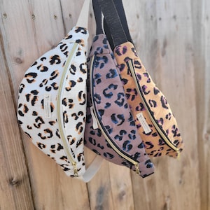 Puede incluir: Tres riñoneras de estampado de leopardo con cremalleras doradas. Las riñoneras están colgadas de una superficie de madera. Las riñoneras son de color blanco, marrón y naranja.