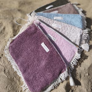 Può includere: Una collezione di cinque pochette rettangolari con cerniera in spugna testurizzata nei toni del verde scuro, marrone, azzurro, beige e viola. Ogni pochette presenta un bordo con frange bianche, una nappa bianca e un'etichetta con L&A CREATION.