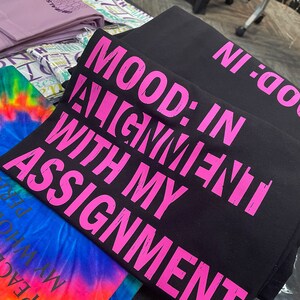 May include: Black t-shirt with pink text that reads "MOOD: IN ALIGNMENT WITH MY ASSIGNMENT".