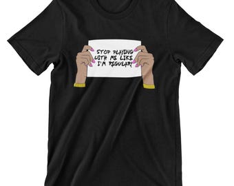 May include: Black t-shirt featuring a white sign that reads "Stop playing with me like I'm regular!" The sign is being held by two hands with pink nail polish.