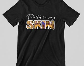 May include: Black t-shirt featuring the text "Pretty in my SKIN" in white script. The word "SKIN" is filled with a colourful, patterned design in yellow, purple, blue, and brown. The shirt has short sleeves and a crew neck.