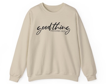 Könnte beinhalten: Ein beigefarbenes Sweatshirt mit Rundhalsausschnitt und dem schwarzen Schriftzug "good thing" in Schreibschrift, wobei der kleinere Text "PROVERBS 18:22" direkt darunter in einer klaren, serifenlosen Schrift mittig auf der Brust steht.