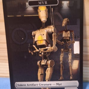 May include: A trading card featuring two robot figures. The card has the word "MYR" at the top and "Token Artifact Creature - Myr" at the bottom. The card is a proxy and not for sale.