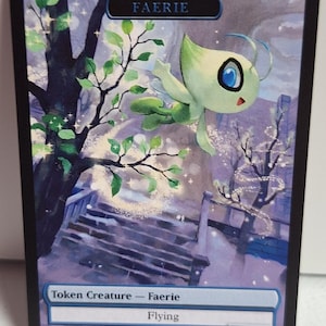 May include: A fantasy card featuring a green and white faerie creature with large blue eyes, flying near a tree. The card has the text "FAERIE", "Token Creature - Faerie", "Flying", and "1/1". The background shows a snowy landscape.