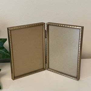 May include: A hinged, double-sided photo frame with a gold-colored metal frame. The frame has a decorative border and is open, revealing the interior backing. The frame is designed to hold two photos and is suitable for displaying on a table or shelf.