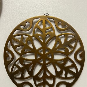 May include: A round, bronze-colored metal wall hanging with a floral design. The intricate pattern features a series of curved lines and petal-like shapes, creating a decorative and openwork effect. A small loop is at the top for hanging.