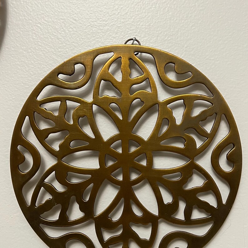 May include: A round, bronze-colored metal wall hanging with a floral design. The intricate pattern features a series of curved lines and petal-like shapes, creating a decorative and openwork effect. A small loop is at the top for hanging.