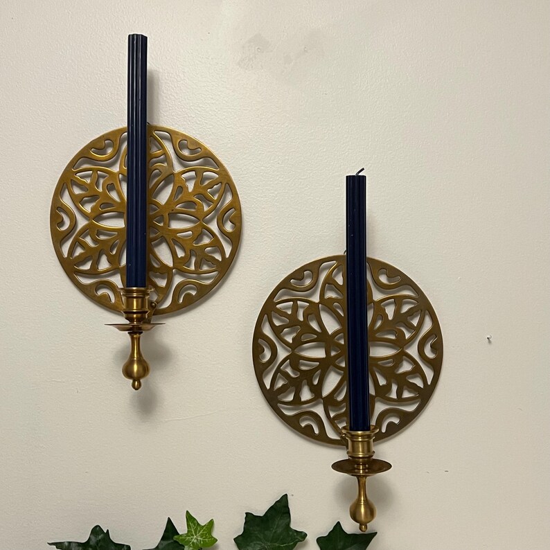 May include: Two brass wall sconces with intricate circular designs. Each sconce holds a tall, navy blue candle. The sconces have a decorative, ornate pattern and a classic, elegant style. The sconces are mounted on a white wall.