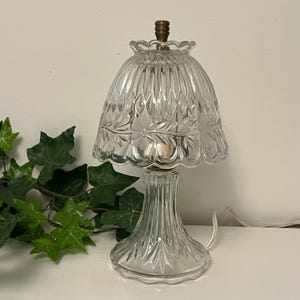 May include: A clear glass table lamp with a scalloped shade and base. The shade features a leaf pattern and the base has vertical ridges. The lamp has a brass-colored top and a white cord.