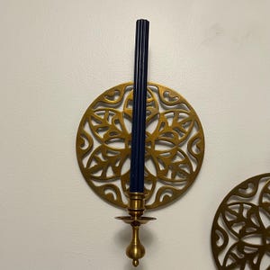 May include: A brass candle holder with a dark blue candle. The candle holder has a decorative, circular design with a floral pattern. The candle is approximately 10 inches tall. The candle holder is mounted on a white wall.