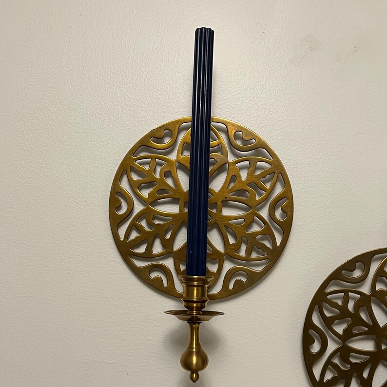 May include: A brass candle holder with a dark blue candle. The candle holder has a decorative, circular design with a floral pattern. The candle is approximately 10 inches tall. The candle holder is mounted on a white wall.