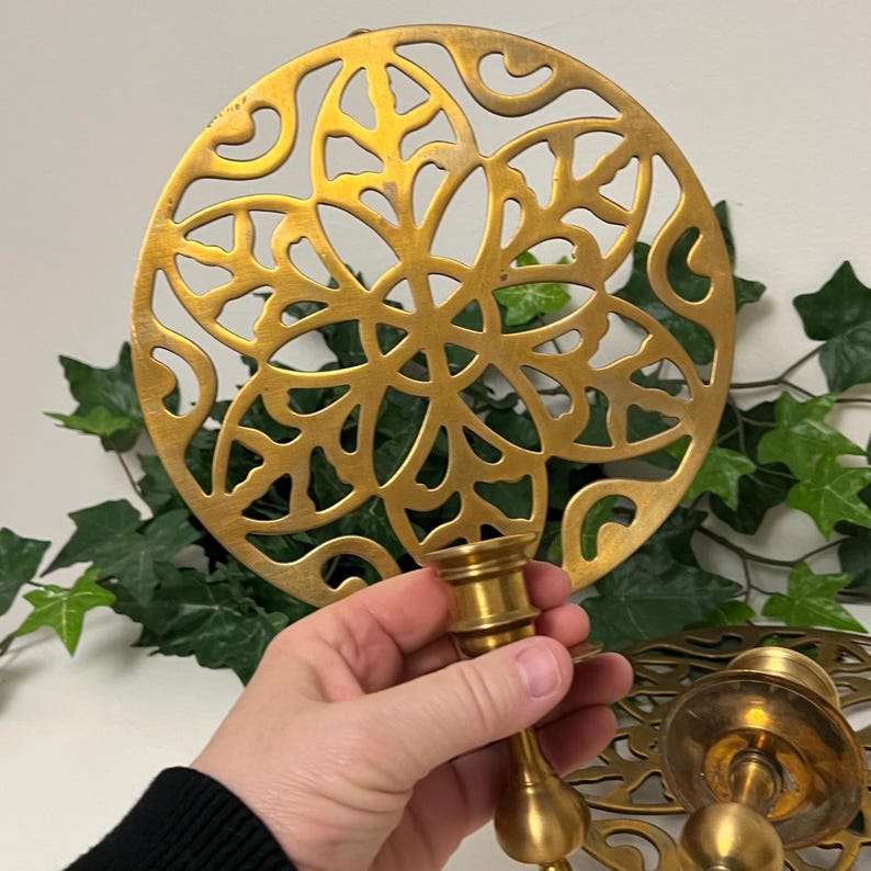 May include: A gold-colored metal candle holder with a decorative, openwork design. The candle holder is held in a hand, and a matching trivet is in the background. The design is circular with a floral pattern.