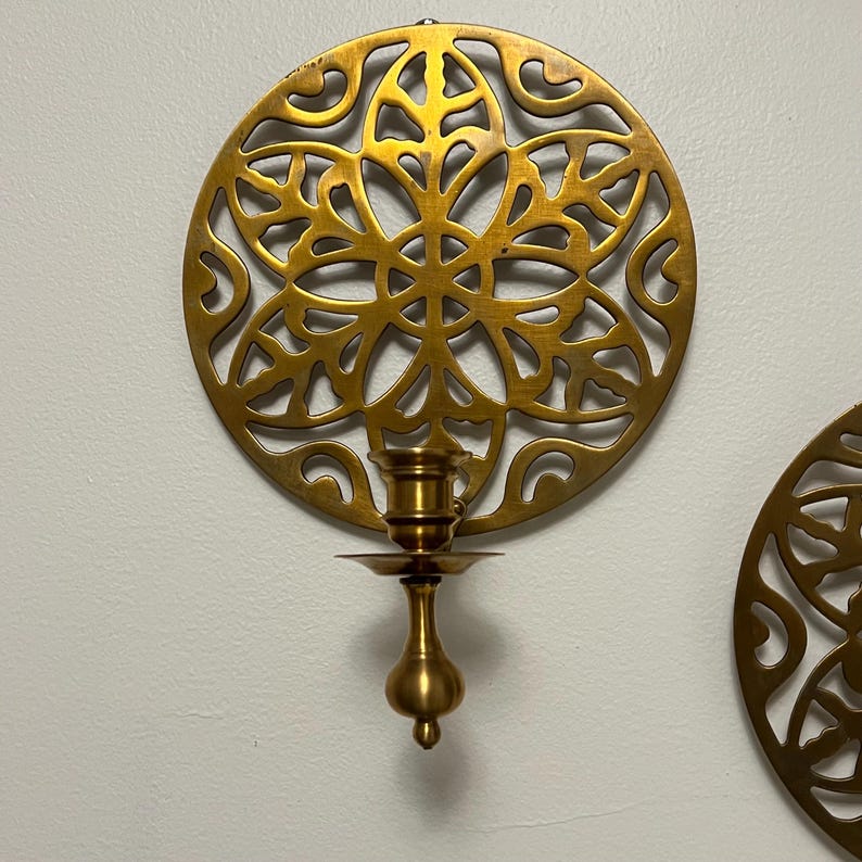 May include: A gold-colored wall sconce with a circular, openwork design. The sconce features a candle holder with a small dish and a decorative base. The intricate pattern is reminiscent of floral or geometric shapes. The sconce is mounted on a white wall.