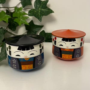 May include: Two decorative Japanese Kokeshi doll-shaped containers. One has a black lid, the other an orange lid. Both have white faces with smiling expressions, and are painted with floral and geometric patterns in blue, orange, and purple.