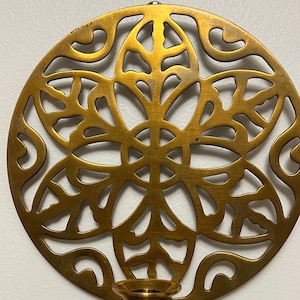 May include: A round, golden-colored metal wall sconce with an intricate, openwork floral design. The sconce features a small cup for holding a candle. The design is symmetrical and detailed, with a vintage aesthetic.