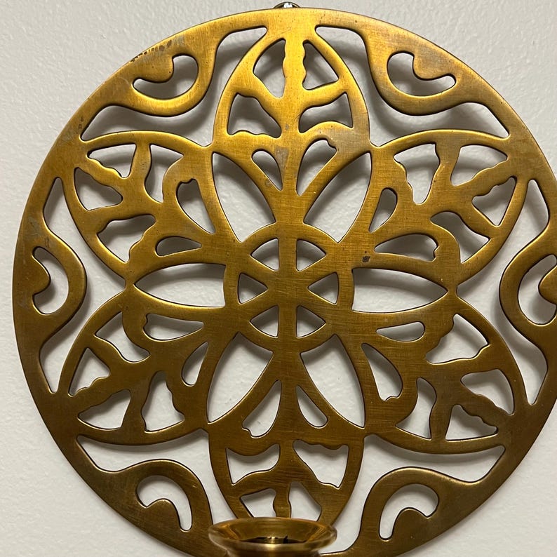 May include: A round, golden-colored metal wall sconce with an intricate, openwork floral design. The sconce features a small cup for holding a candle. The design is symmetrical and detailed, with a vintage aesthetic.