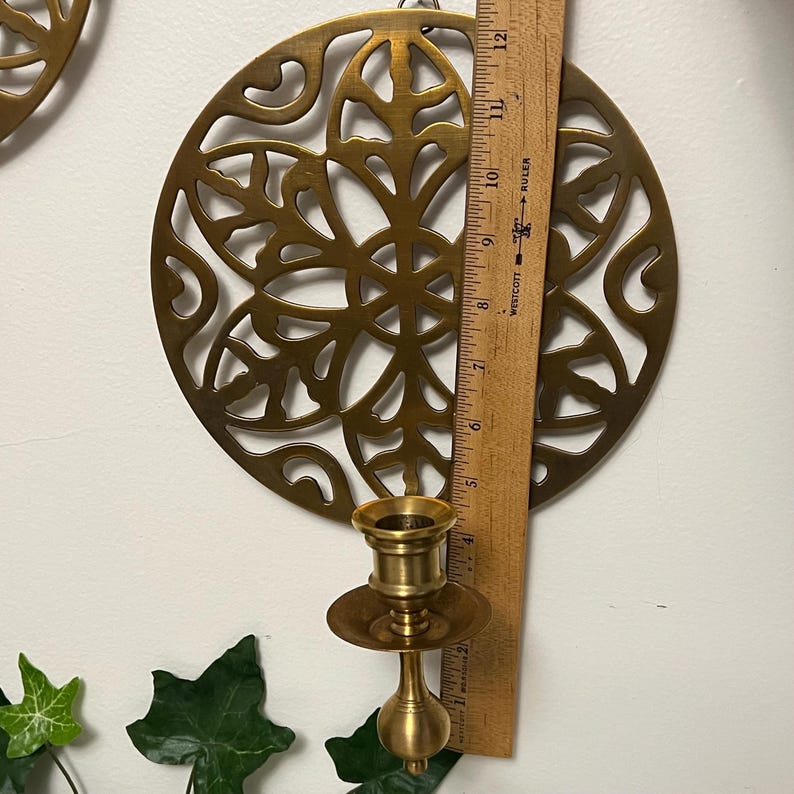 May include: A brass candle holder with a decorative circular design. The candle holder is approximately 4 inches tall. A wooden ruler is placed next to the candle holder, showing the measurement in inches. The wall decor is a vintage style.