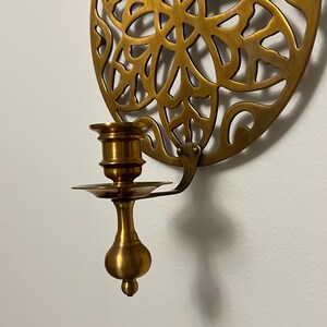 May include: A brass wall-mounted candle holder. The candle holder has a decorative, circular backplate with an openwork design. The candle cup and base are brass, with a small dish to catch wax. The holder is mounted on a white wall.
