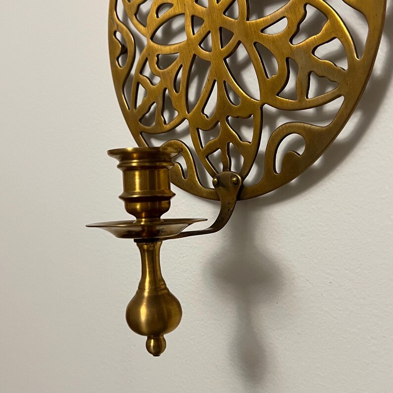 May include: A brass wall-mounted candle holder. The candle holder has a decorative, circular backplate with an openwork design. The candle cup and base are brass, with a small dish to catch wax. The holder is mounted on a white wall.