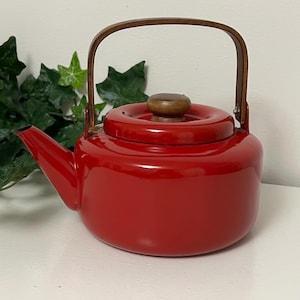 May include: A bright red enamel teapot featuring a rounded body, a short spout, and a matching red lid with a small, round, dark brown wooden knob. The teapot has a tall, arched, dark brown wooden handle attached with silver metal hardware.