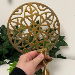 May include: A gold-colored metal candle holder with a round base and a decorative, openwork design. The candle holder is held in front of a matching gold-colored, circular trivet with a floral pattern. Green ivy is in the background.