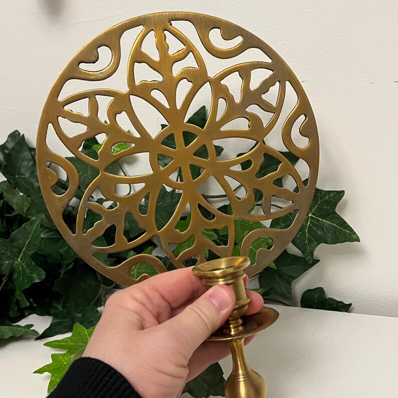 May include: A gold-colored metal candle holder with a round base and a decorative, openwork design. The candle holder is held in front of a matching gold-colored, circular trivet with a floral pattern. Green ivy is in the background.