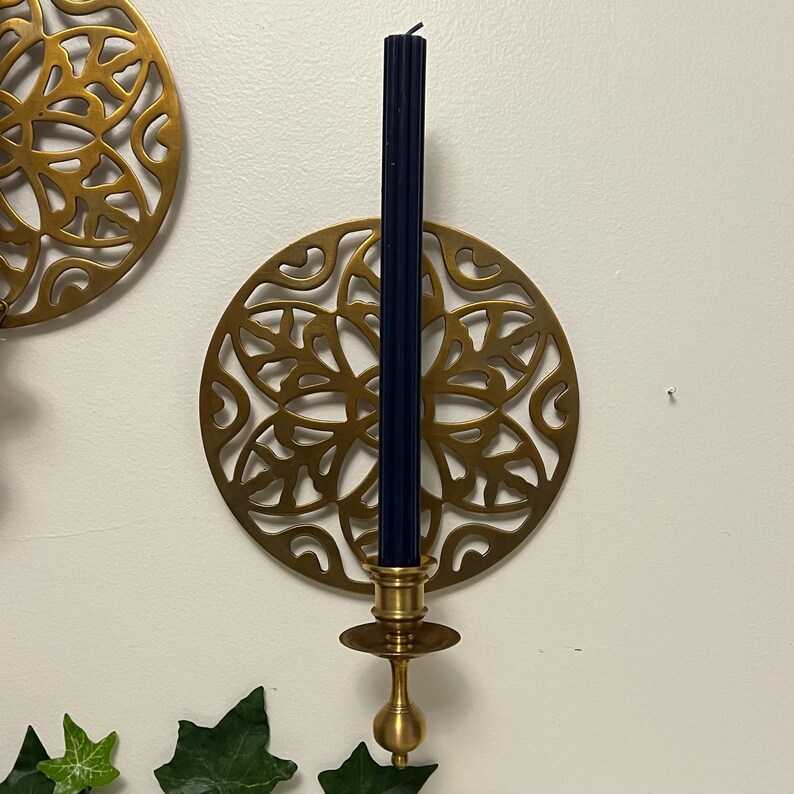 May include: A brass candle holder with a navy blue candle. The candle holder has a decorative, circular design. The candle is tall and slender. The candle holder is mounted on a white wall.